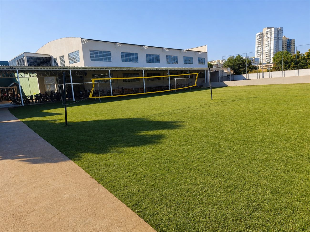 Campo gramado com rede de vôlei e o ginásio ao fundo
